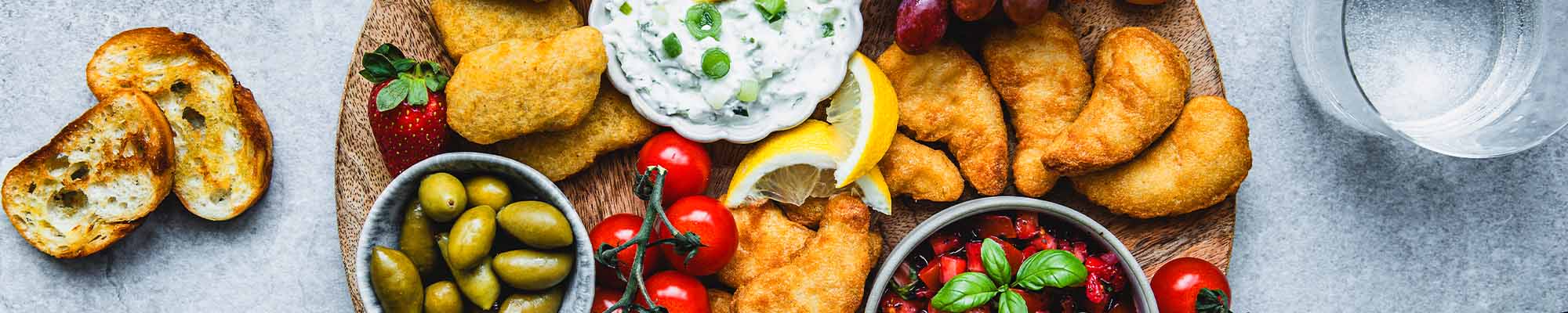 Buntes Sommer-Snack-Board mit Chicken Nuggets, Dippers, Tomaten-Erdbeer-Salsa, Fetacreme, Tomaten, Oliven, Trauben und Baguette